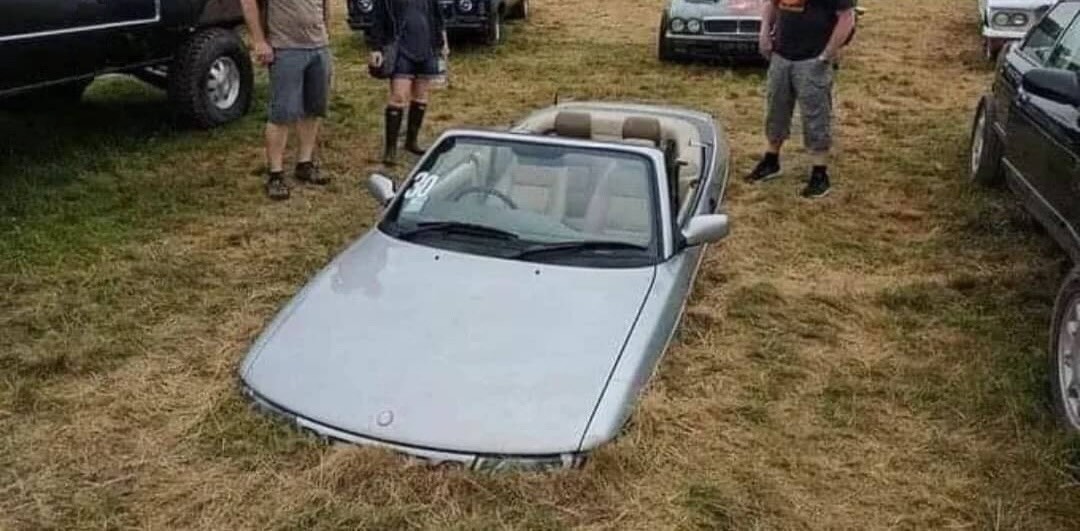 The bottom halves of three people looking at a silver convertible buried half deep in the ground. Portions of five other cars are in the background.