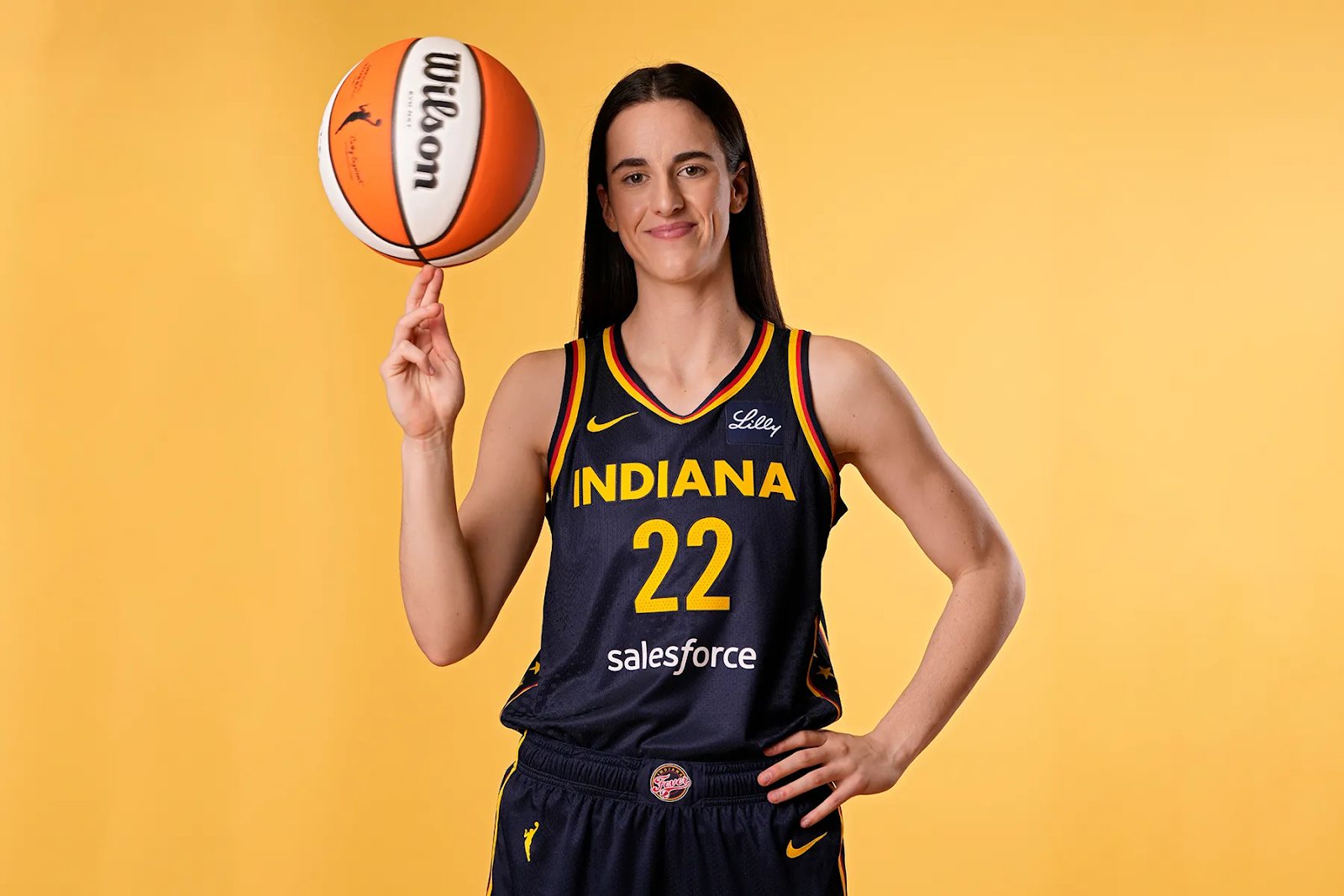 Caitlin Clark in Indiana Fever uniform #22, smiling and spinning a basketball on two fingers of her right hand.  The uniform has logos for sponsors Nike, Eli Lilly, and SalesForce.