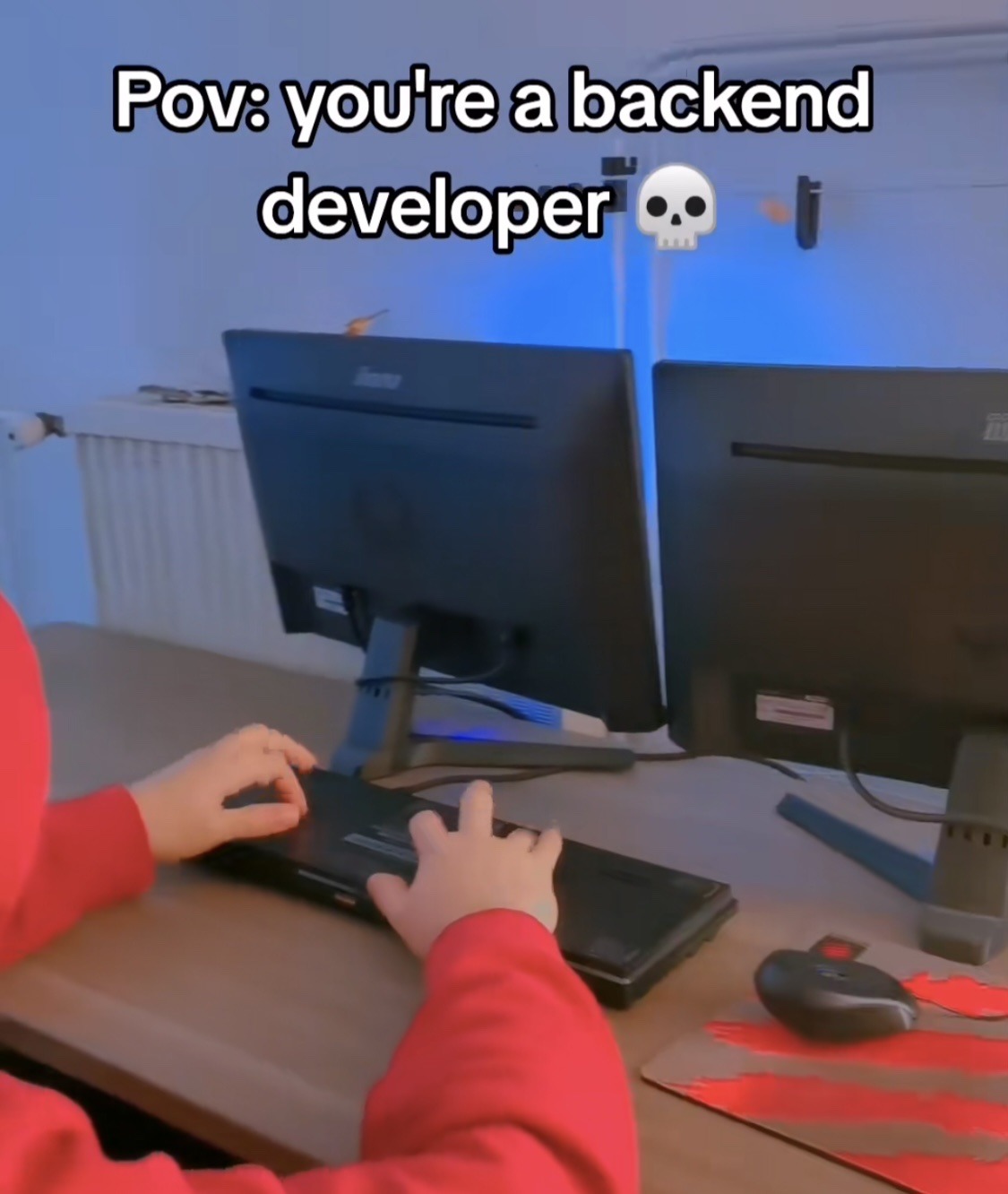 A person in a red hoodie, mostly obscured, looking at the back of a computer monitor. Their hands are on an upside-down keyboard. The mouse is upside-down.