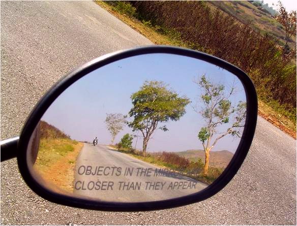 A rear-view mirror seen through the passenger-side window of an auto, with road and brush in the background. On the mirror is the standard caption, in all caps, 'Objects in the mirror closer than they appear'